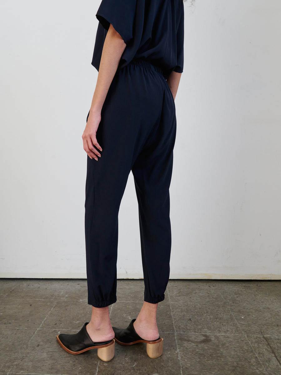 A person stands sideways in a room with a concrete floor and white wall, dressed in a dark blue ensemble featuring Zero + Maria Cornejo's Gabi Trousers, which come with hidden pockets. The outfit includes loose-fitting pants crafted from stretch silk charmeuse and is paired with black clogs that have wooden heels.