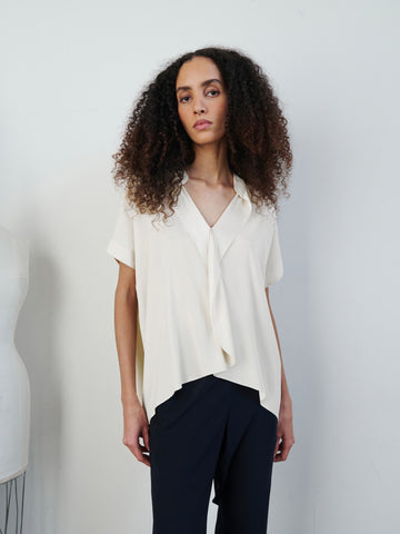 A person with long, curly hair stands against a plain white background, wearing the Fin Gaban Shirt by Zero + Maria Cornejo— a short-sleeved, loose-fitting cream blouse featuring draped ruffle detail— and black pants. A dress form is partially visible on the left side.