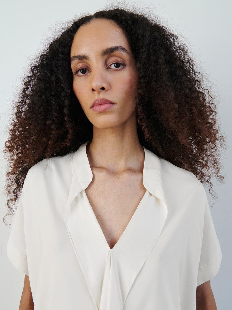 A person with long, curly hair and wearing the Fin Gaban Shirt by Zero + Maria Cornejo, featuring a white silk charmeuse fabric and a stand collar, gazes directly at the camera. The background is plain and light-colored.