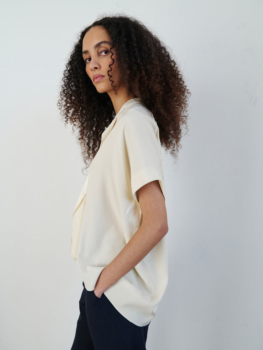 A person with curly hair stands against a plain backdrop, wearing the Fin Gaban Shirt by Zero + Maria Cornejo—a light-colored silk charmeuse top featuring draped ruffle detail—and dark pants, exuding confidence as they look at the camera.