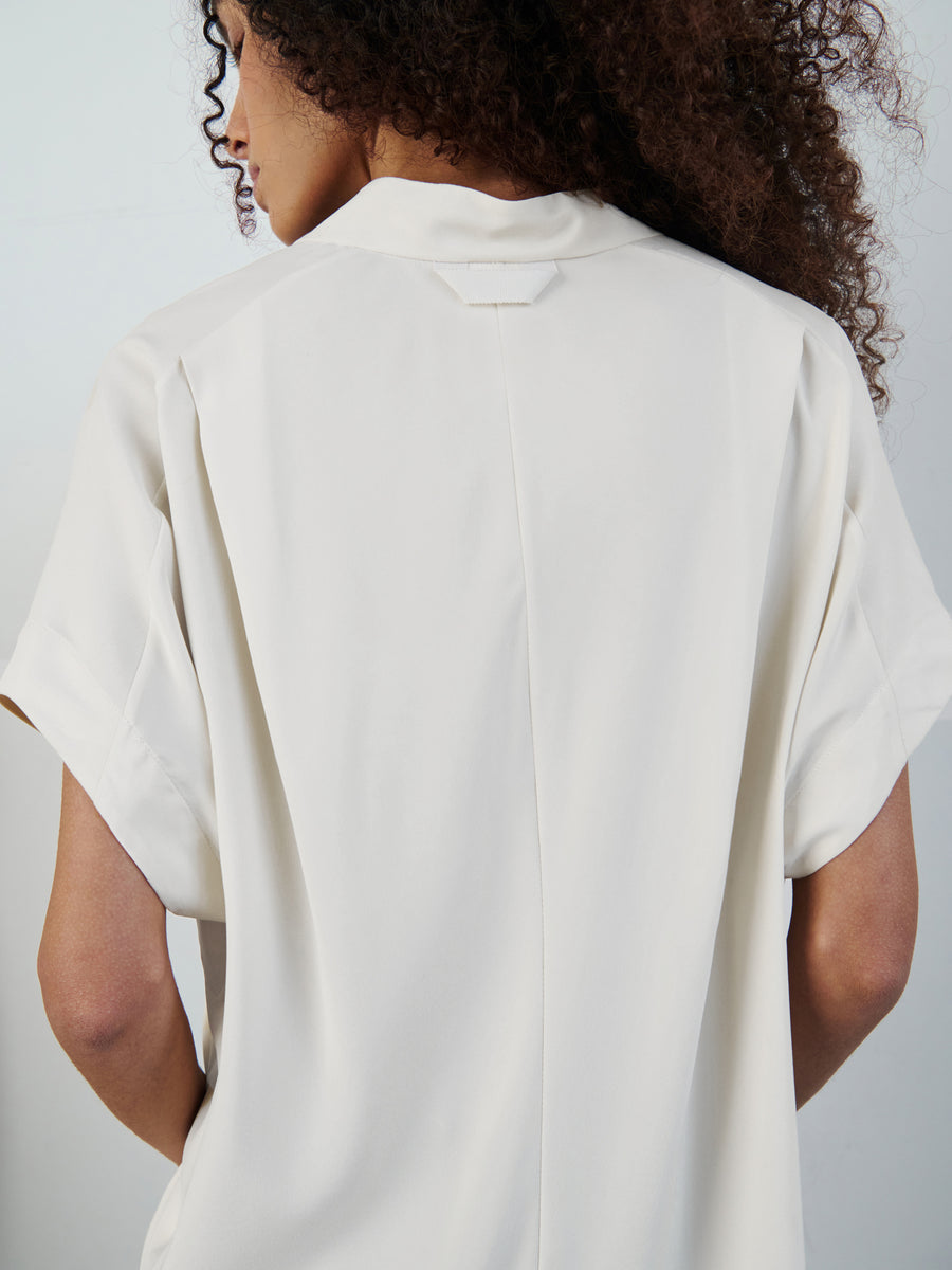 A person with curly brown hair facing away, wearing the off-white Fin Gaban Shirt by Zero + Maria Cornejo, featuring rolled-up sleeves. The background is plain and minimalistic.