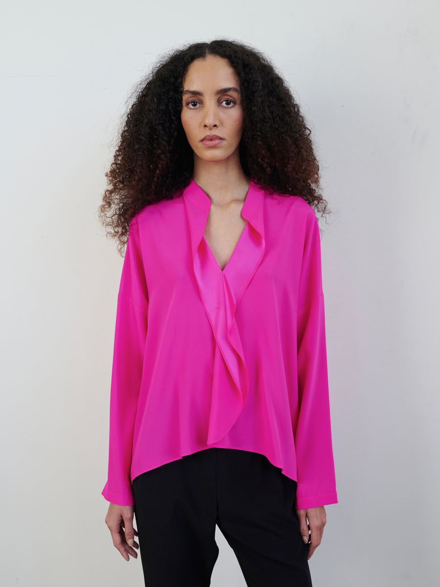 A person with curly hair is wearing a vibrant pink Long-Sleeved Fin Gaban Shirt by Zero + Maria Cornejo, which features a draped ruffle neckline, paired with black pants. The background is plain and light-colored.