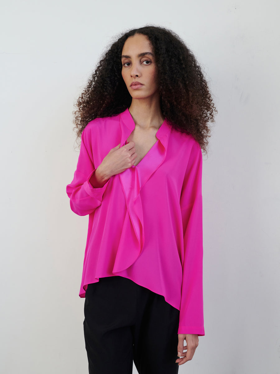 A person with long, curly hair is wearing a Long-Sleeved Fin Gaban Shirt by Zero + Maria Cornejo in bright pink, featuring a draped ruffle neckline and paired with dark pants. They stand against a plain white background, looking directly at the camera with a neutral expression.