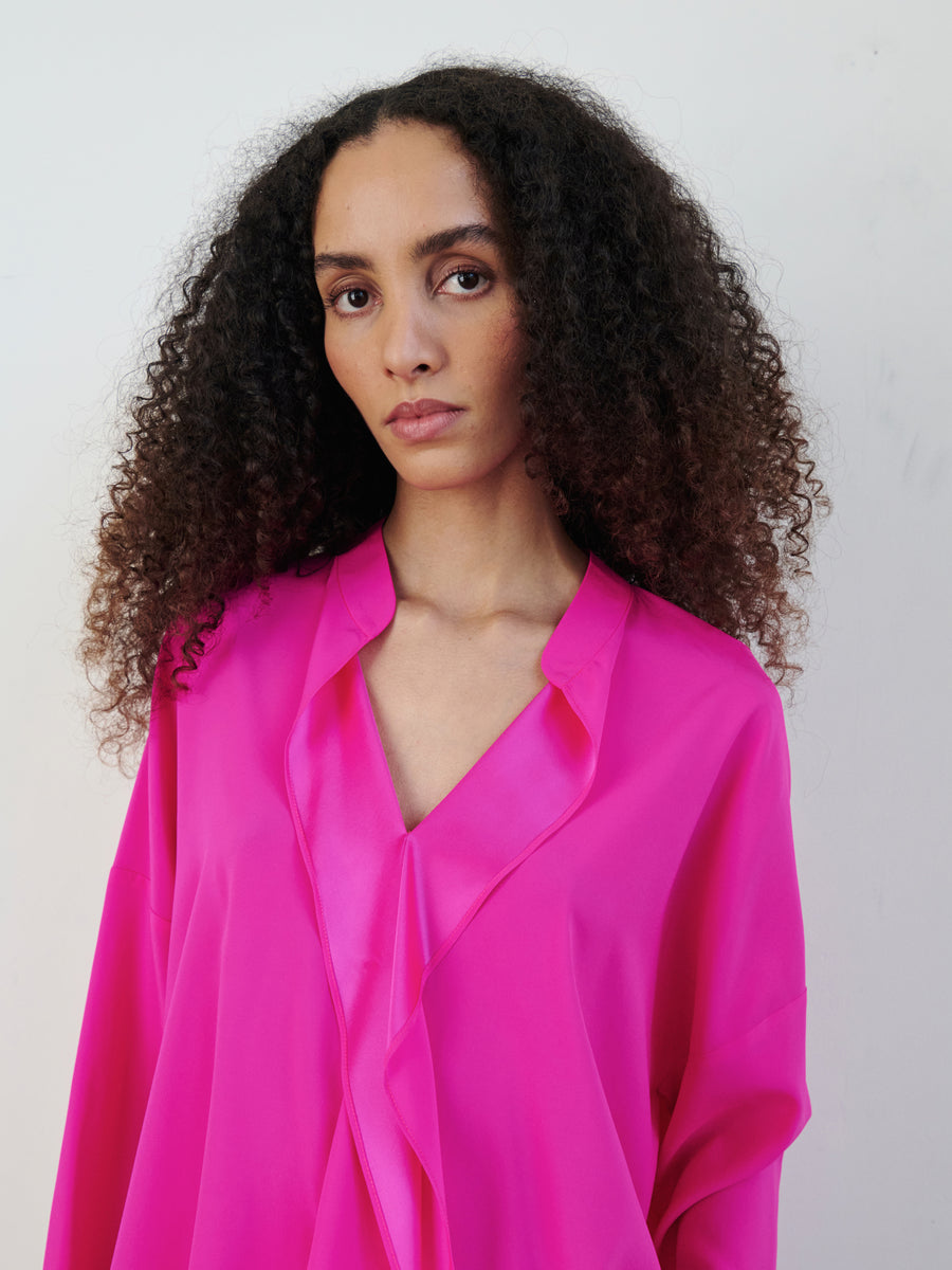 A person with long curly hair is wearing a vibrant pink Long-Sleeved Fin Gaban Shirt by Zero + Maria Cornejo, featuring a draped ruffle neckline. They stand against a plain light background, looking directly at the camera with a neutral expression.