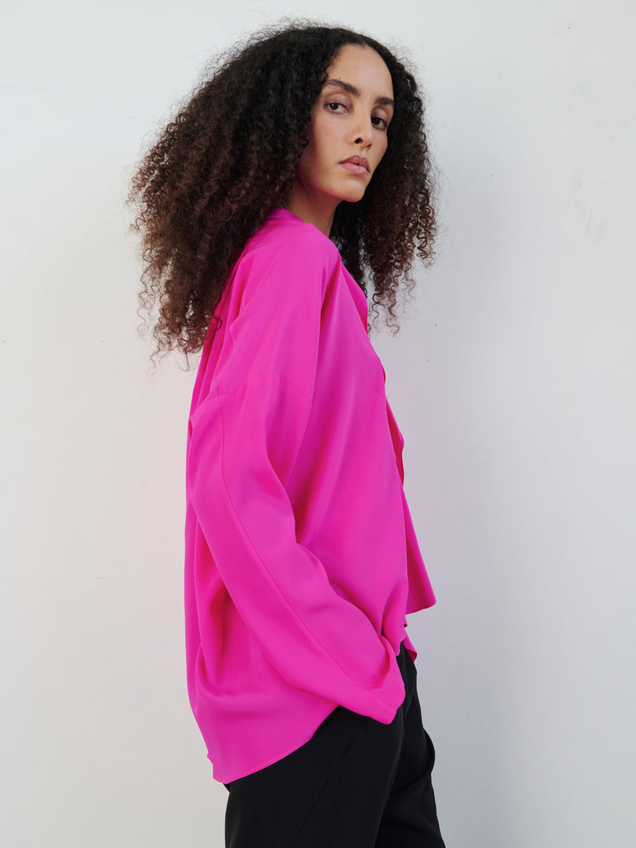 A person with curly hair is wearing a vibrant pink Long-Sleeved Fin Gaban Shirt by Zero + Maria Cornejo and black pants. They are standing against a plain white background, their gaze thoughtfully drifting to the side.