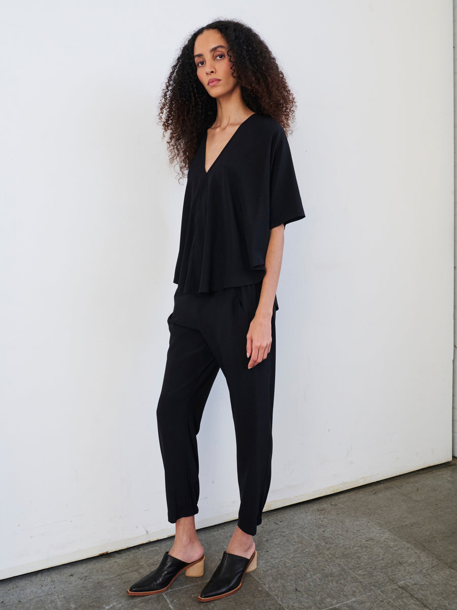 A person with long curly hair stands against a white wall, wearing a loose-fitting black top and Zero + Maria Cornejo's Gabi Trouser made from stretch silk charmeuse. They have a confident pose and are looking towards the camera, their ensemble completed by black slip-on shoes.