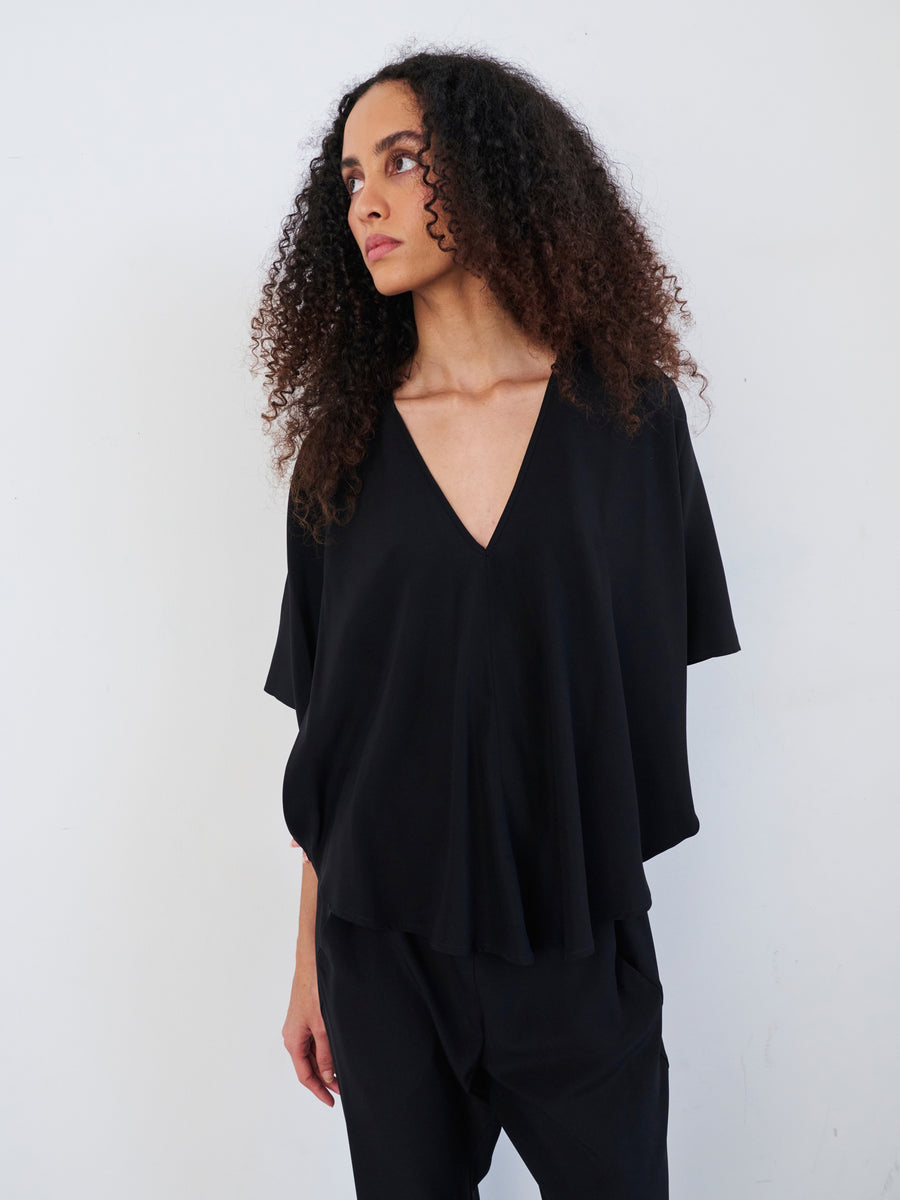 A person with curly hair, dressed in a draped silhouette featuring the Circle Top by Zero + Maria Cornejo and matching pants, looks to the side against a plain white background.