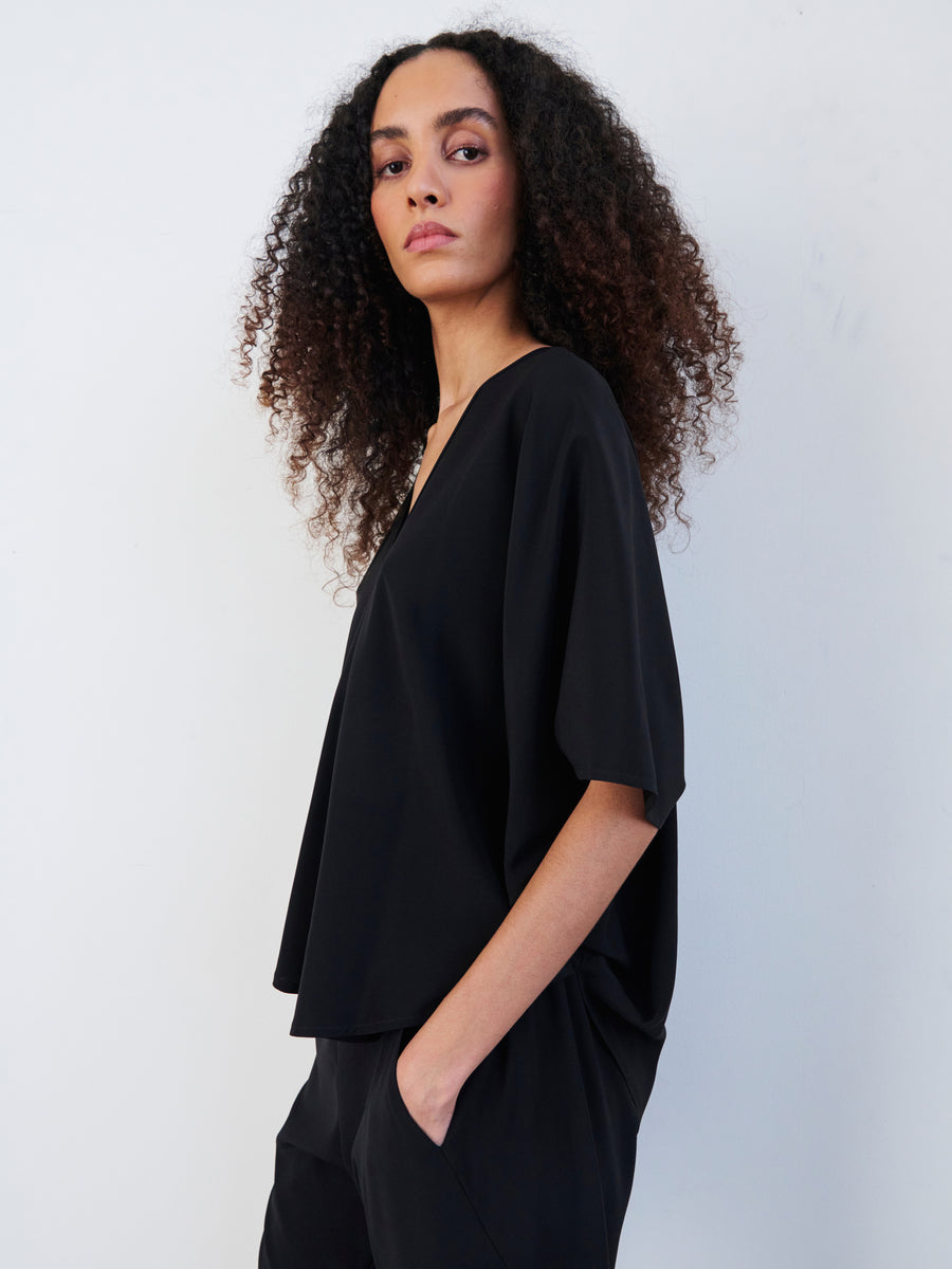 A person with curly hair is confidently modeling the Zero + Maria Cornejo Circle Top, a loose-fitting black outfit with a draped silhouette, against a plain white background.