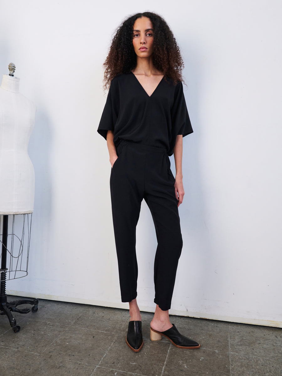 A person with long, curly hair stands confidently in a minimalist room wearing the Gabi Trouser from Zero + Maria Cornejo. They pair this elegant piece with black mules and pose next to a white dress form on the wooden floor, effortlessly showcasing the sophistication of stretch silk charmeuse.