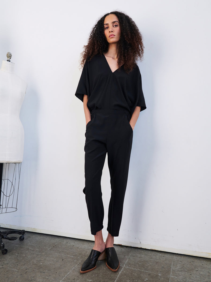 A person with long, curly hair is elegantly dressed in the Gabi Trouser from Zero + Maria Cornejo, a black V-neck jumpsuit crafted from stretch silk charmeuse. They complete the look with sleek black shoes, standing in a bright room beside a white mannequin form. Their outfit exudes sophistication, featuring discreetly integrated hidden pockets.