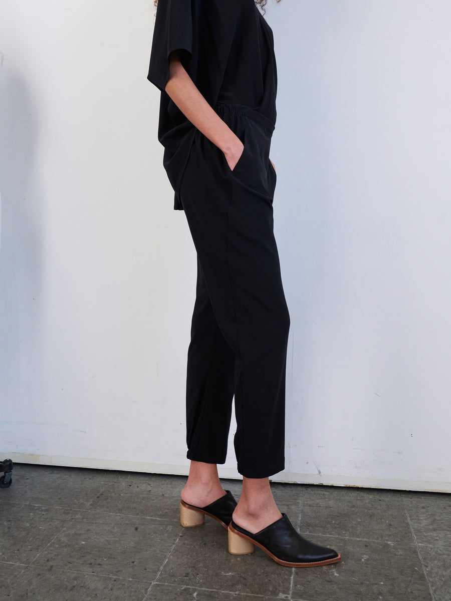 A person stands with their hands in their pockets, wearing a loose black outfit featuring stylish Gabi Trousers from Zero + Maria Cornejo and brown heeled mules. The background is a white wall.