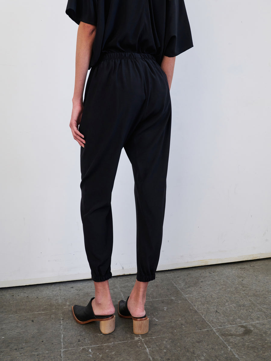 A person dressed in a black ensemble consists of the stretch silk charmeuse Gabi Trousers by Zero + Maria Cornejo, showcasing loose-fitting pants and short sleeves, stands facing a white wall. They are wearing black high-heel clogs with wooden soles and have one hand by their side, appreciating the convenience of hidden pockets.