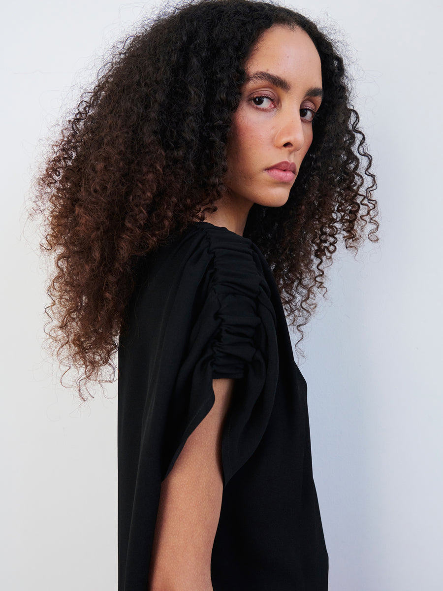 A person with curly hair, wearing a black Ruched Muscle Top by Zero + Maria Cornejo made from woven viscose, stands against a plain background, looking over their shoulder.