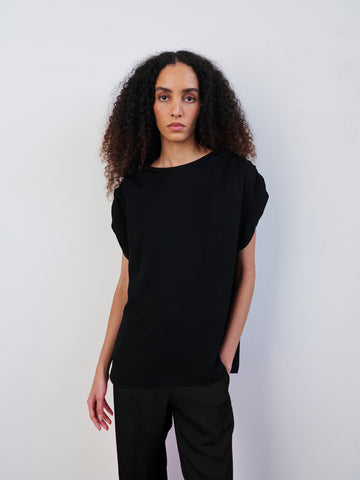A person with long curly hair is wearing the Ruched Muscle Top from Zero + Maria Cornejo paired with woven viscose pants, both in a loose-fitting black style. They stand against a plain white backdrop with their hands by their sides and a neutral expression. The outfit is made from sustainably-sourced materials, emphasizing an eco-friendly drape.