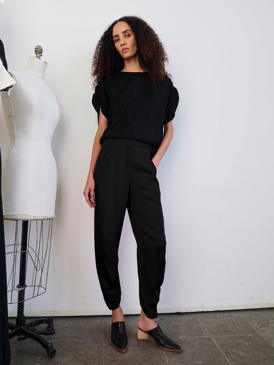 A person with curly hair stands against a wall in a minimalist room, dressed in the Takeo Pant by Zero + Maria Cornejo. They're wearing a black top and woven viscose black pants, standing beside a dress form with their hands relaxed at their sides, embodying the essence of sustainably-sourced fashion elements.