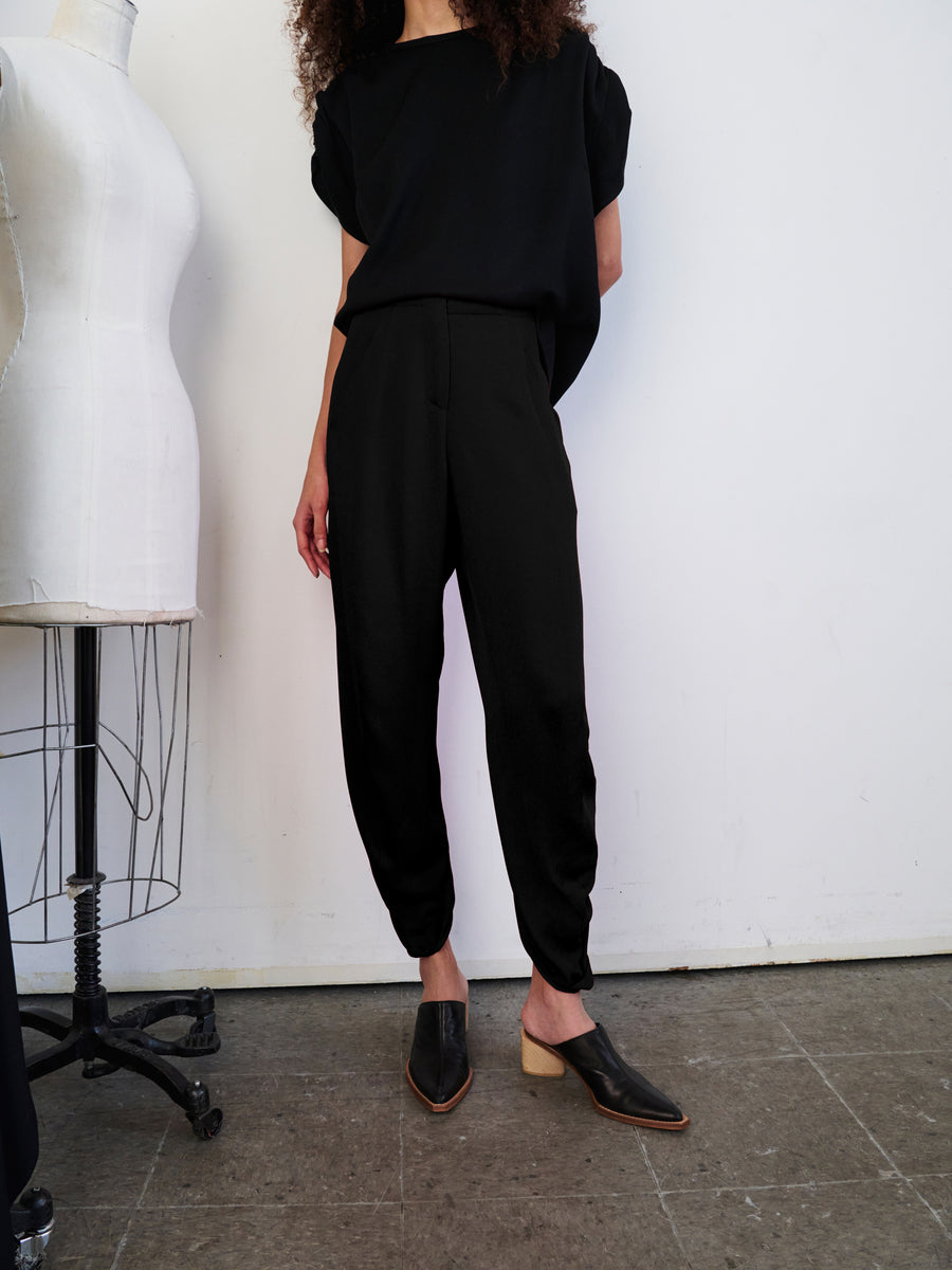 Standing in a minimal studio near a dress form, a person with long curly hair showcases a draped silhouette. They are wearing a loose black top along with the Takeo Pant from Zero + Maria Cornejo, crafted from sustainably-sourced viscose. The outfit is completed with black shoes featuring a tan heel against the plain white background and gray floor.