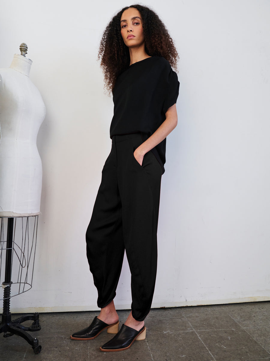 A person with curly hair stands confidently in a black outfit and slip-on shoes, their hands casually nestled in the pockets of the sustainably-sourced Takeo Pant by Zero + Maria Cornejo. Beside them, a dress mannequin complements the scene against a plain white wall.