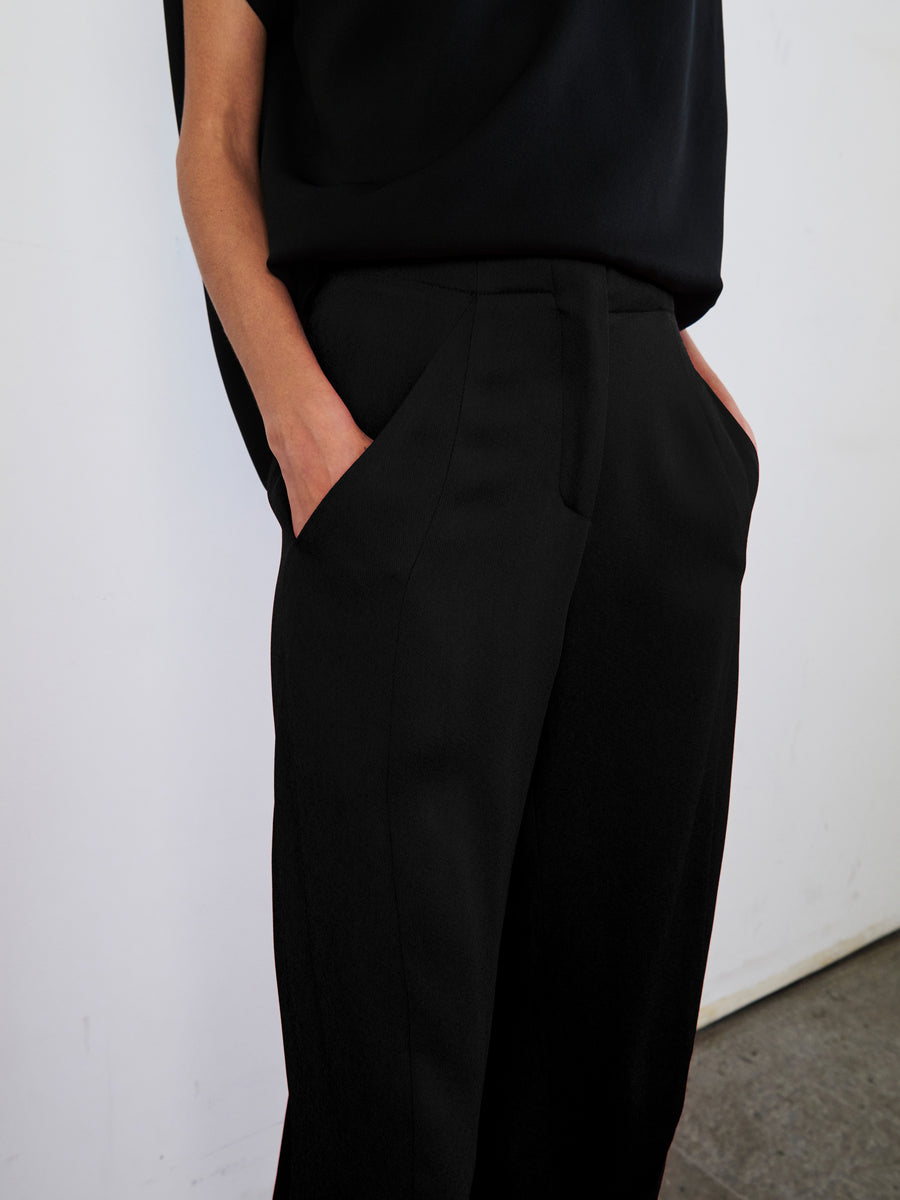 Against a white wall, a person models the Zero + Maria Cornejo black sleeveless top paired with the Takeo Pant, made from sustainably-sourced viscose. Their hands rest casually in their pockets, highlighting the chic draped silhouette of the outfit. The photograph elegantly captures their ensemble from shoulder to mid-thigh.