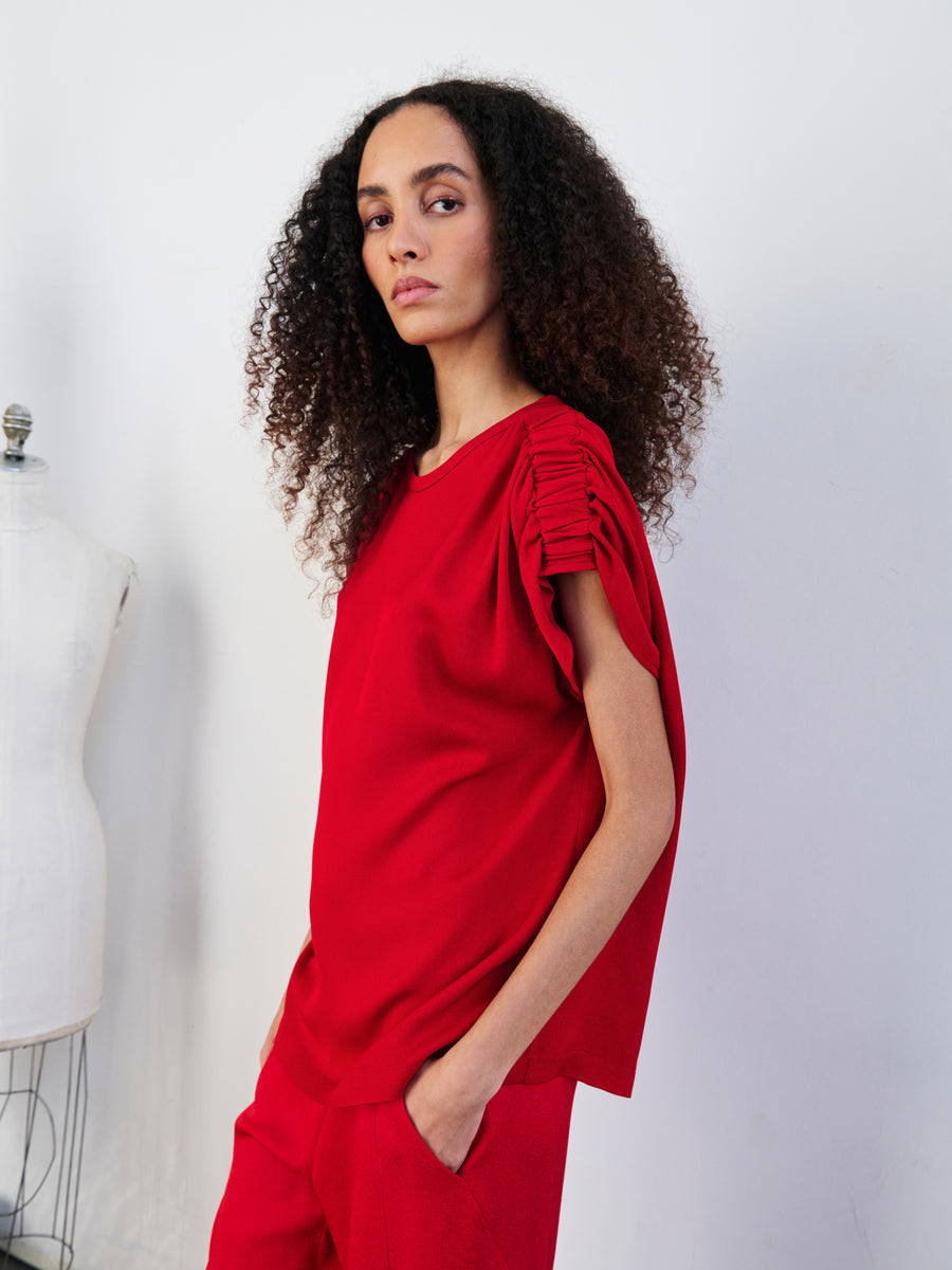 A person with long, curly hair poses in a red Ruched Muscle Top from Zero + Maria Cornejo, featuring ruched shoulder details. They stand with one hand in a pocket and maintain a neutral expression against a plain backdrop, beside a dress form.