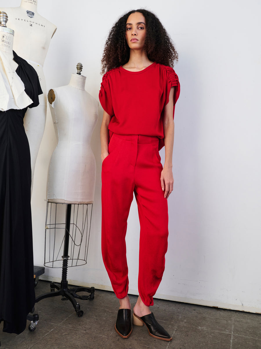 A person with curly hair stands confidently in a vibrant red woven viscose outfit, wearing the high-waisted Takeo Pant and matching short-sleeve top. They are near two mannequins in a minimalist fashion studio that highlights the eco drape fabric from Zero + Maria Cornejo's sustainably-sourced collection.