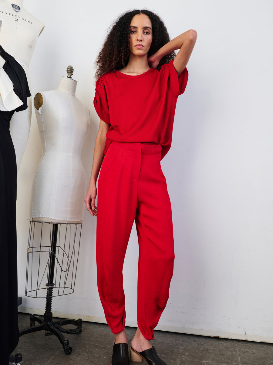 A person wearing a vibrant red Takeo Pant from Zero + Maria Cornejo poses confidently in a studio setting, standing between two dress forms. The white walls and minimal background emphasize the boldness of their outfit, highlighting the draped silhouette crafted from sustainably-sourced viscose.