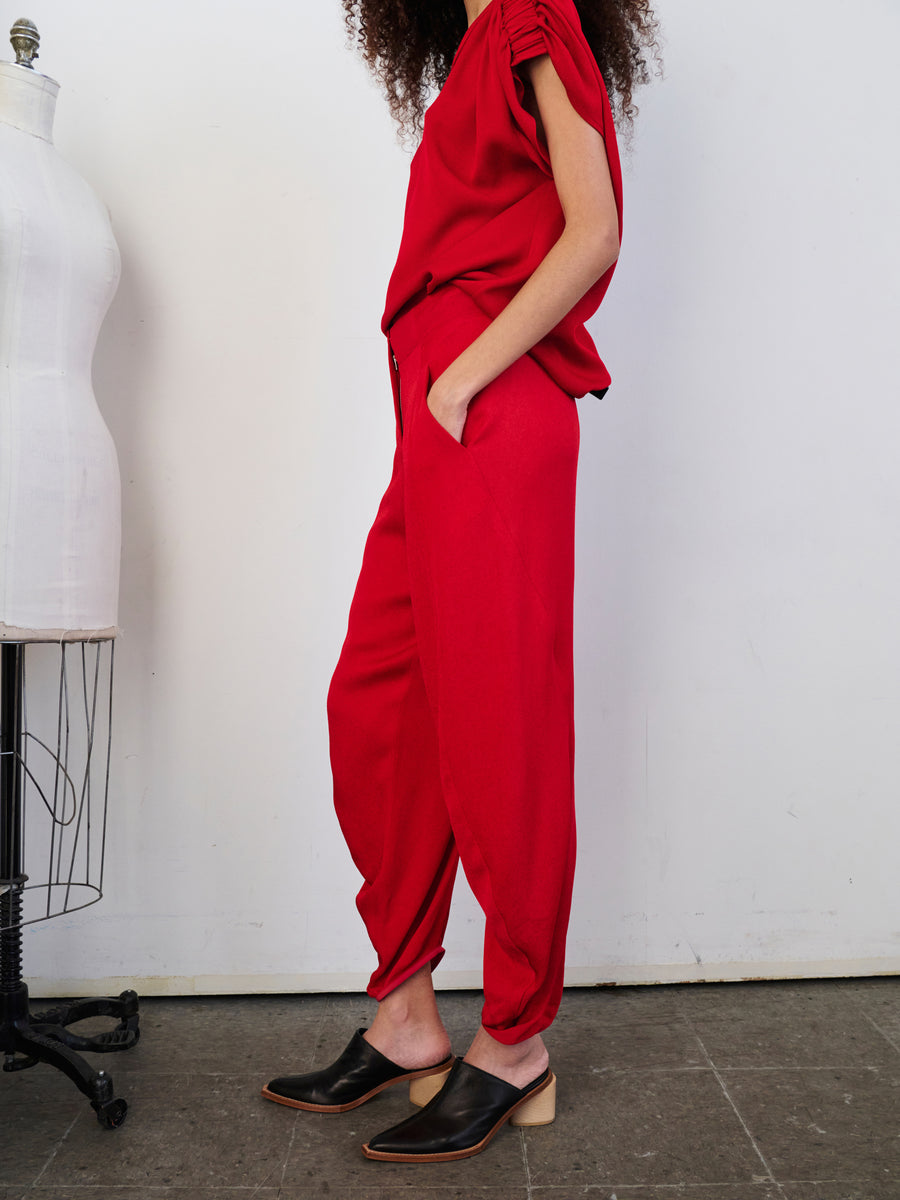 A person stands next to a dress form, wearing the stylish red Takeo Pant by Zero + Maria Cornejo, crafted from sustainably-sourced fabric. They pair it with a sleeveless top and black slip-on shoes. The background is a plain wall.