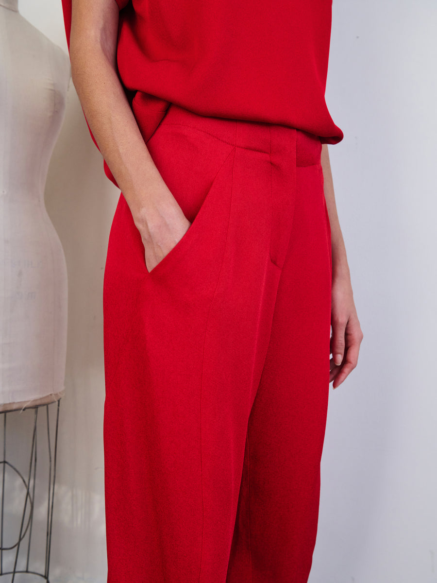 A person dressed in the vibrant red Takeo Pant, crafted from woven viscose with eco drape by Zero + Maria Cornejo, places their hand in the pocket while standing beside a dress form. The plain, light-colored wall in the background accentuates the vibrancy and sustainable sourcing of the clothing.