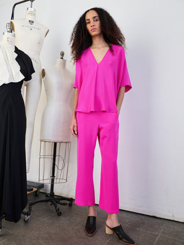 A person with long curly hair stands confidently in a bright pink two-piece outfit featuring the Eko Pant from Zero + Maria Cornejo. They are in a studio with dress mannequins, one of which is draped in a black dress. The person wears black pointed shoes.