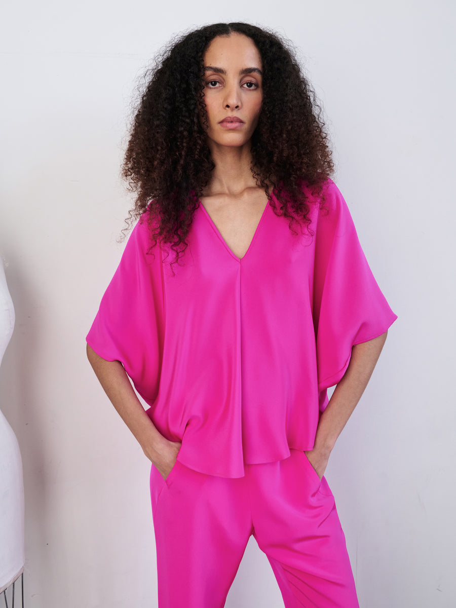 A person with curly hair wears the vibrant pink Circle Top by Zero + Maria Cornejo, along with matching pants, forming a draped silhouette as they stand against a plain backdrop with hands in pockets.