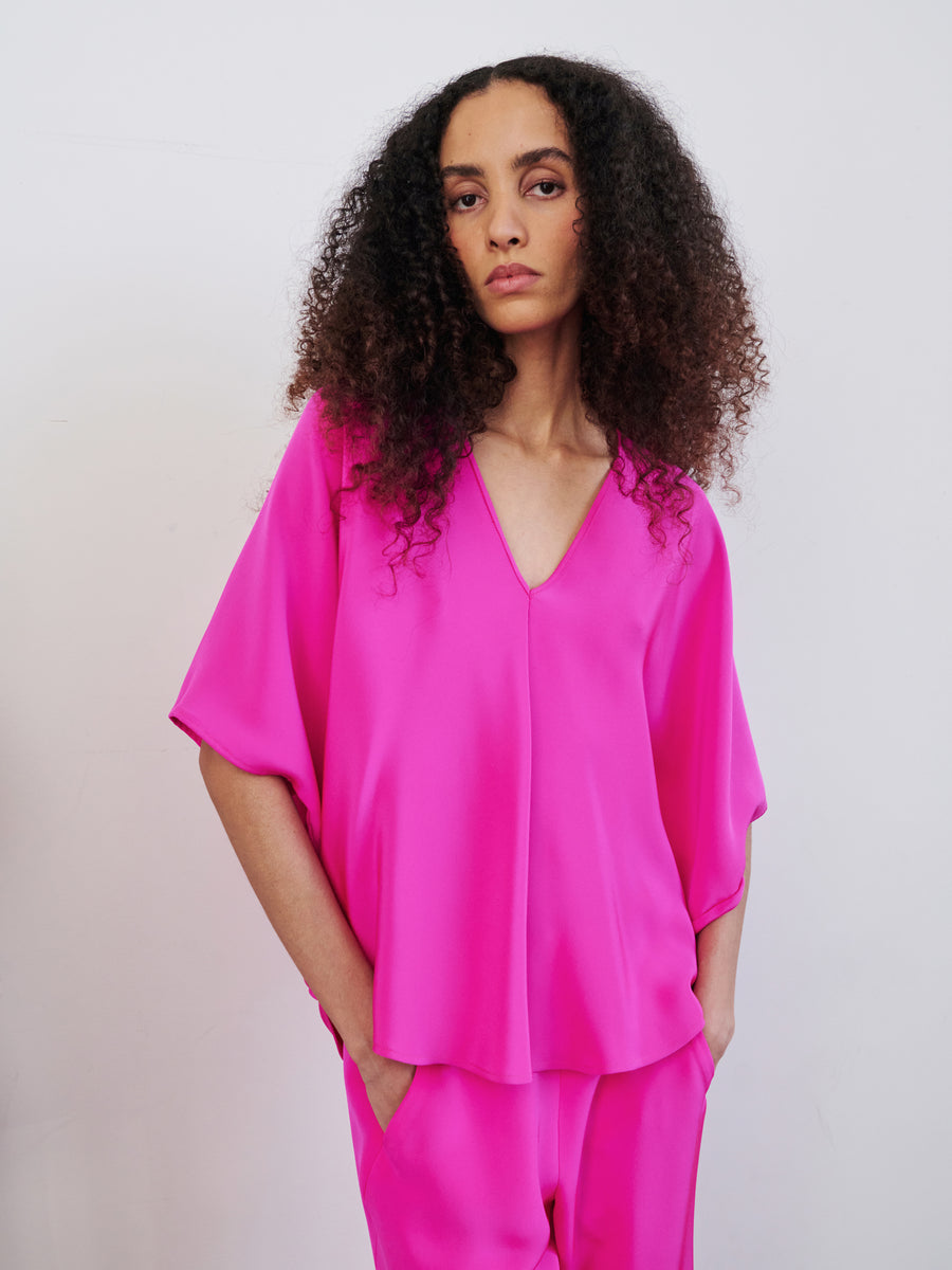 A person with curly hair is wearing the Circle Top by Zero + Maria Cornejo in a vibrant pink silk V-neck style, along with matching pants. The draped silhouette accentuates their confident stance against a plain white background as they gaze assuredly towards the camera.