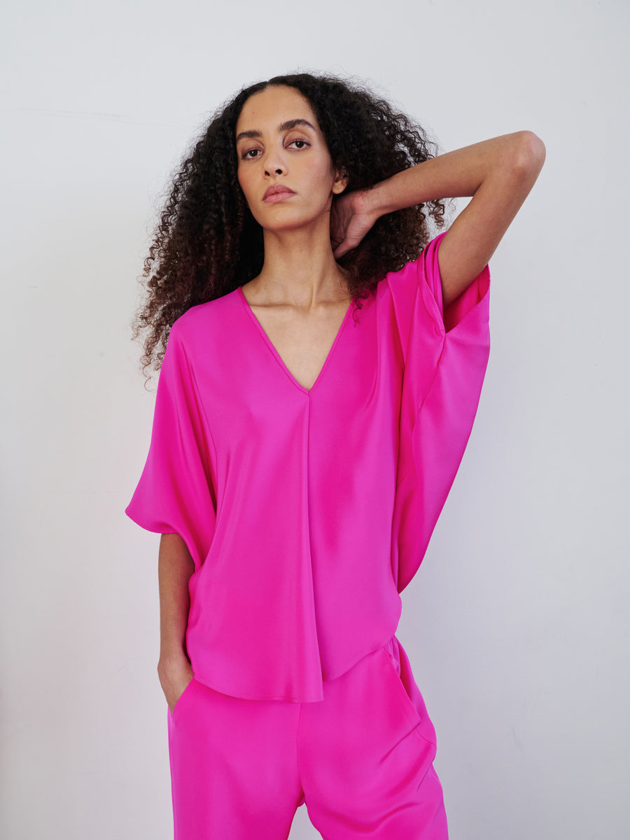 A person with curly hair poses confidently in the vibrant Circle Top by Zero + Maria Cornejo. With one hand in their pocket and the other resting on their head, they exude a relaxed elegance against a plain backdrop.