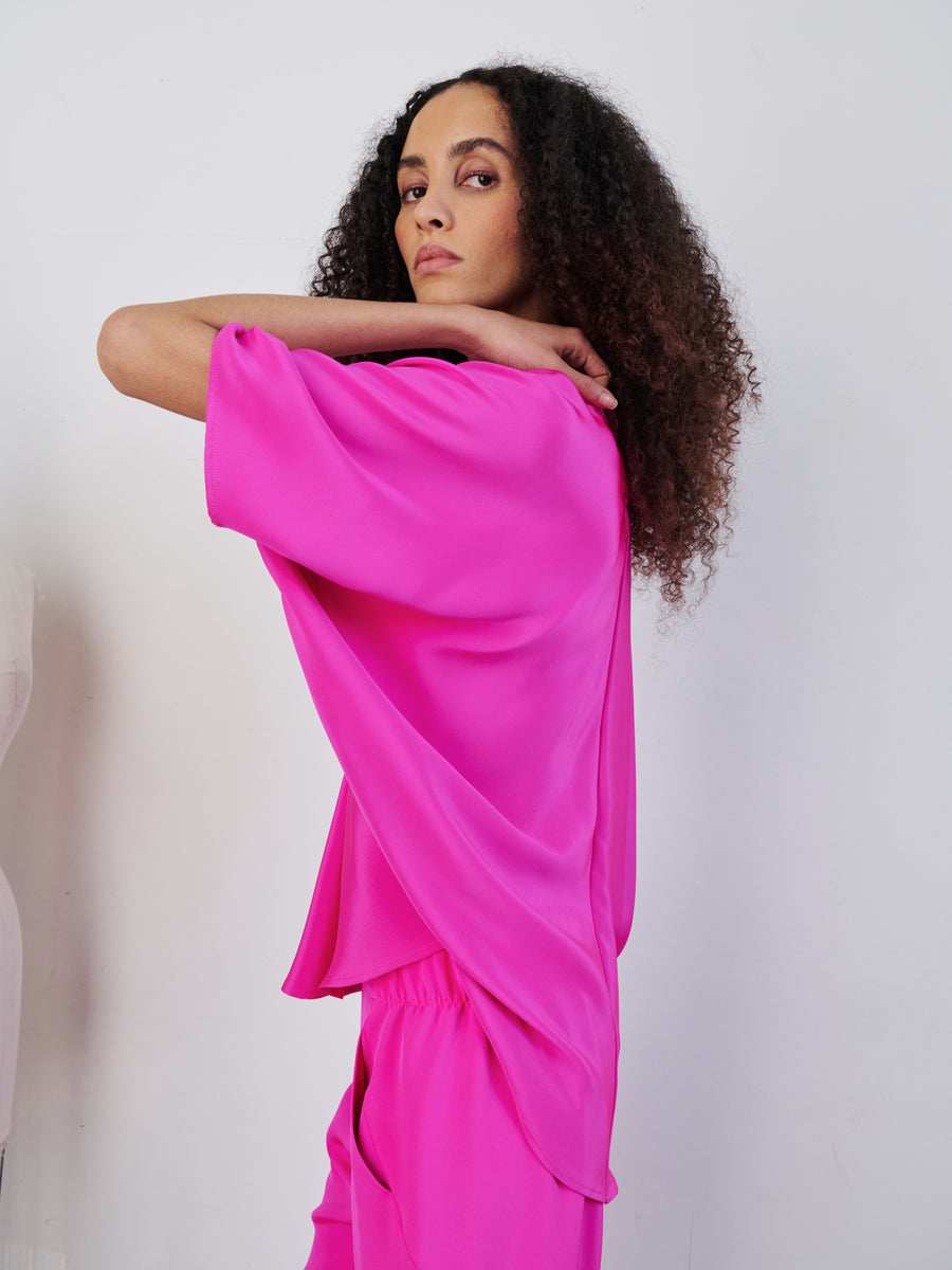 A person with curly hair poses confidently in a bright pink Circle Top from Zero + Maria Cornejo, featuring a v-neck, set against a plain white background. They look over their shoulder with a neutral expression, highlighting the loose-fitting draped silhouette of the top and matching pants.