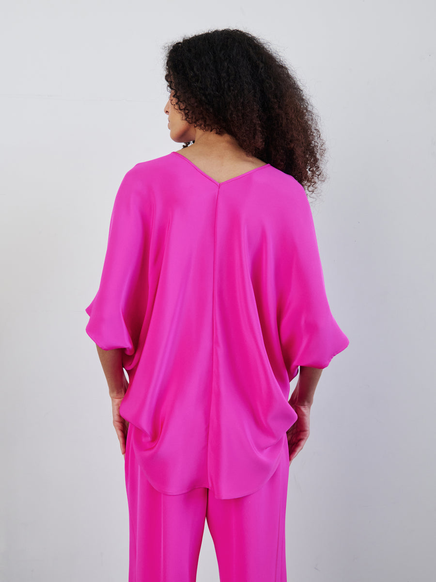 A person with curly hair stands facing away, wearing a loose, flowing bright pink Circle Top and matching pants by Zero + Maria Cornejo. The v-neck design and draped silhouette add an elegant touch against the plain white background, highlighting the vibrant outfit.