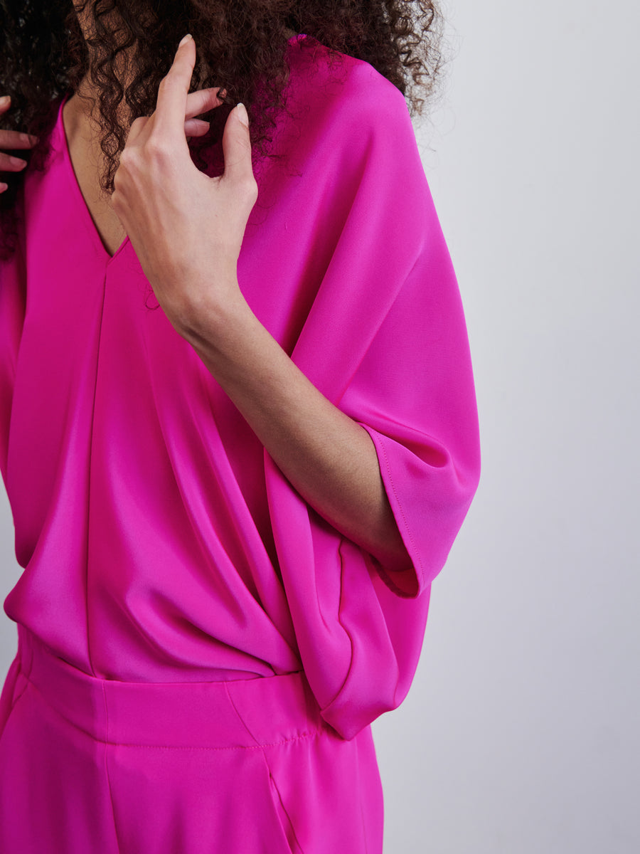 The individual is adorned in a Circle Top from Zero + Maria Cornejo, featuring a vibrant pink silk design with a V-neck and loose sleeves, as they hold their curly hair near their head. The outfit boasts a draped silhouette and is paired perfectly with matching pants.