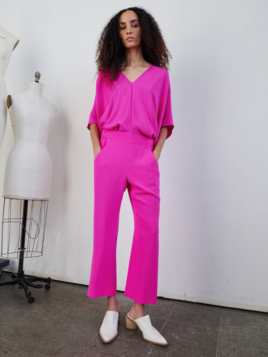 In a minimalist studio with a concrete floor, a person with curly hair stands confidently in a vibrant pink outfit next to a dress form. The ensemble includes a V-neck loose top and matching woven silk Eko Pants by Zero + Maria Cornejo, featuring hidden pockets. They complete the look with white pointed shoes.