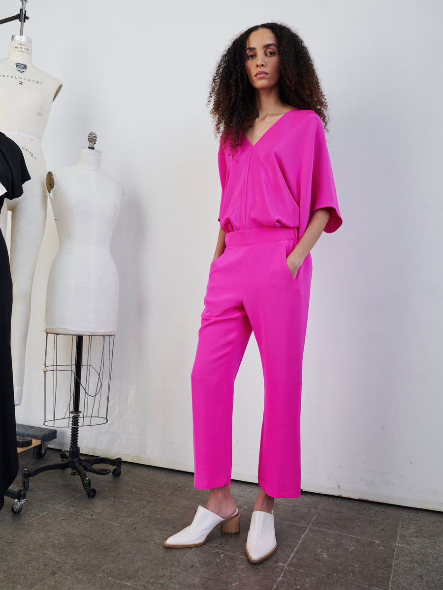 A person with curly hair stands confidently in a bright pink outfit featuring the Eko Pant by Zero + Maria Cornejo, characterized by woven silk trousers with an elastic waist and hidden pockets, paired with white shoes. They are positioned near dress forms in a studio setting.