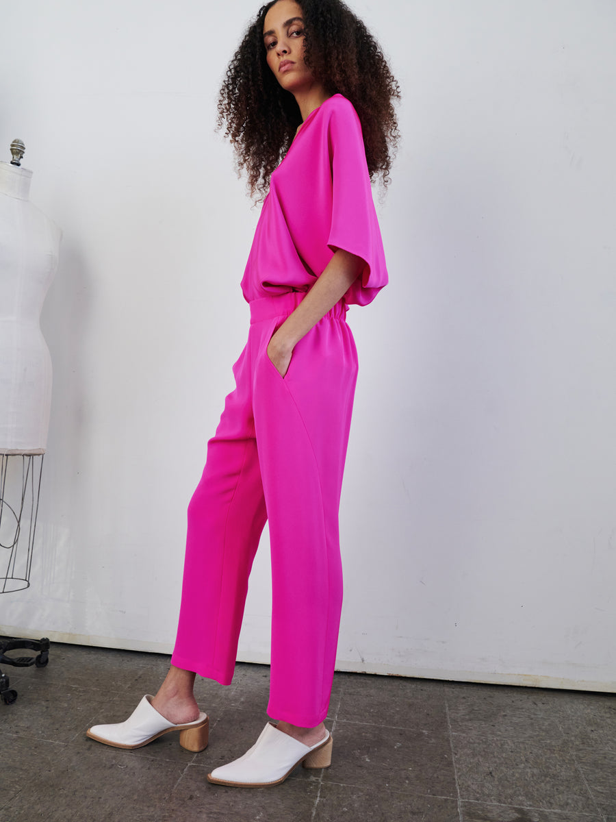 In a minimalist studio, a person with curly hair models the vibrant pink Eko Pant from Zero + Maria Cornejo. Confidently standing beside a dress form, they flaunt their white heeled shoes while hands comfortably nestled in the hidden pockets of their woven silk trousers and matching top.