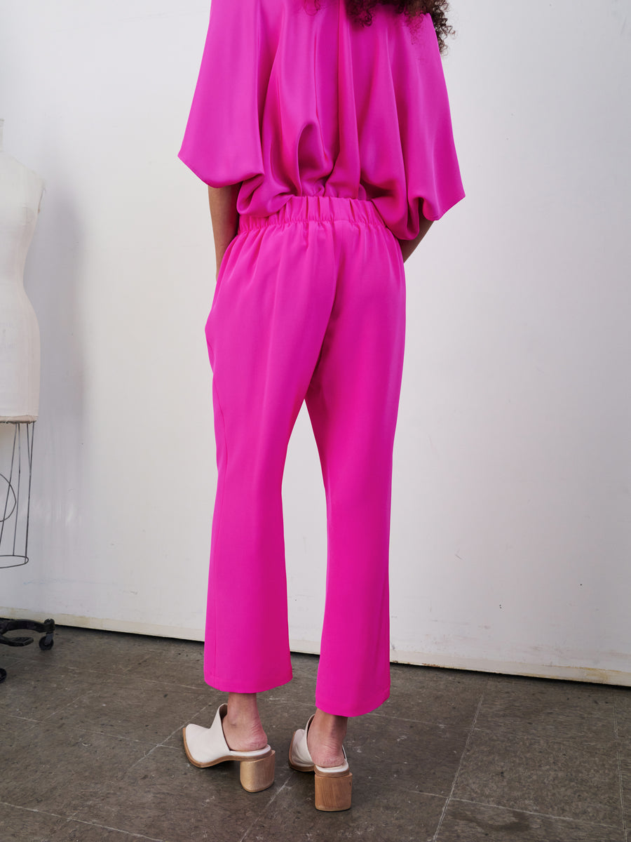 A person stands turned away, spotlighting a vivid pink blouse combined with the Eko Pant by Zero + Maria Cornejo. The trousers are woven silk with an elastic waist, and the ensemble is completed with white heeled shoes. The backdrop features a neutral wall, and on the left, a mannequin is partially visible.