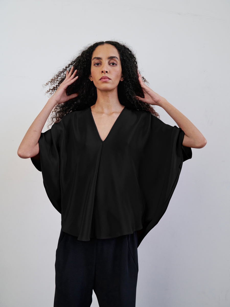 A person with long, curly hair is wearing a loose, black V-neck Circle Top by Zero + Maria Cornejo and black pants while standing against a plain white background. They have their hands raised to their head, fingers gently touching their hair, enhancing the blouse's draped silhouette.