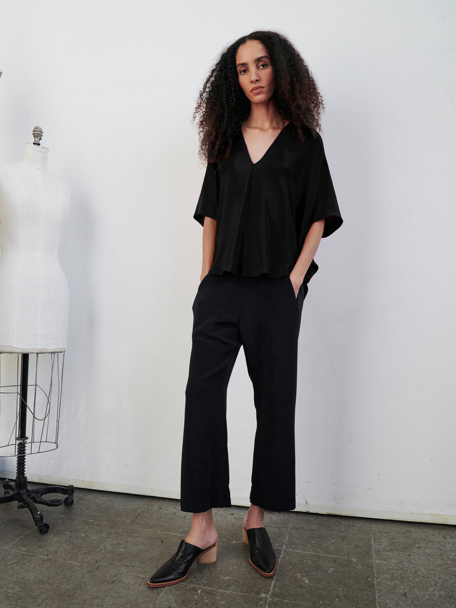 A person with long curly hair stands in a minimalist room, wearing a black V-neck top paired with the Eko Pant by Zero + Maria Cornejo. The woven silk trousers, featuring hidden pockets, complement their relaxed pose beside a dress form mannequin. The concrete floor and white walls highlight the sleek simplicity of the scene.