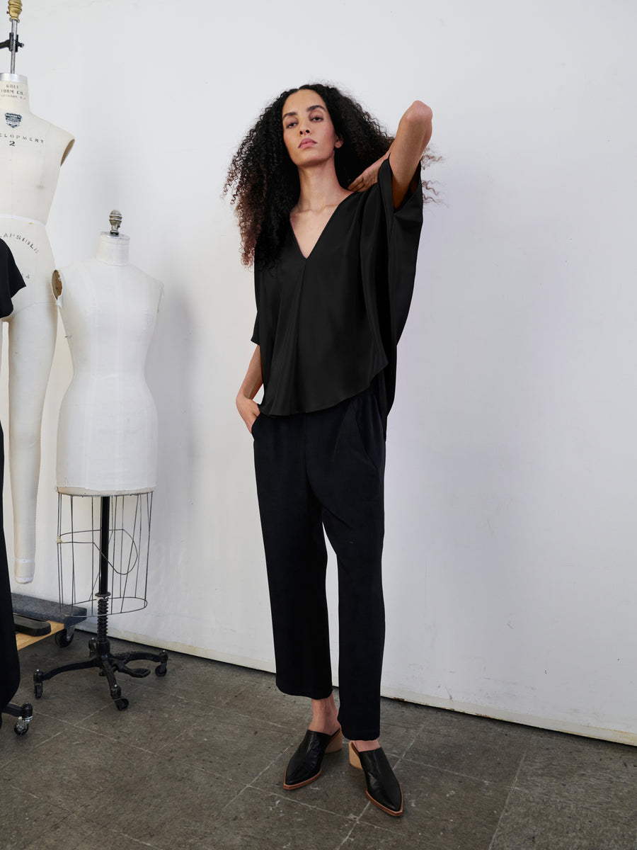 A person with long curly hair stands confidently in a room filled with dress forms, wearing a Circle Top by Zero + Maria Cornejo—a black v-neck silk top—paired with black pants and black pointed shoes. One hand is casually placed in their pocket while the other rests on their hip.