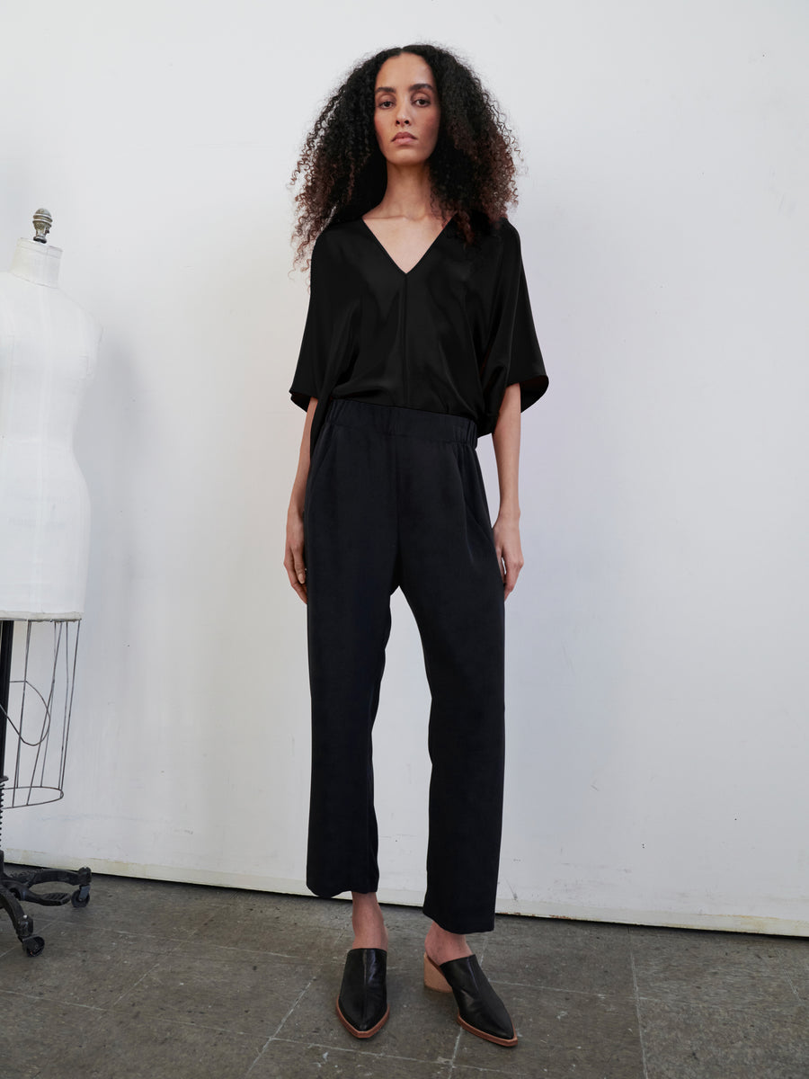 In a minimalist room, a person with long, curly hair stands confidently wearing a loose black V-neck top and the Eko Pant by Zero + Maria Cornejo, featuring an elastic waist and hidden pockets. They complete the look with black slip-on shoes, while a dress form is partially visible to the left.