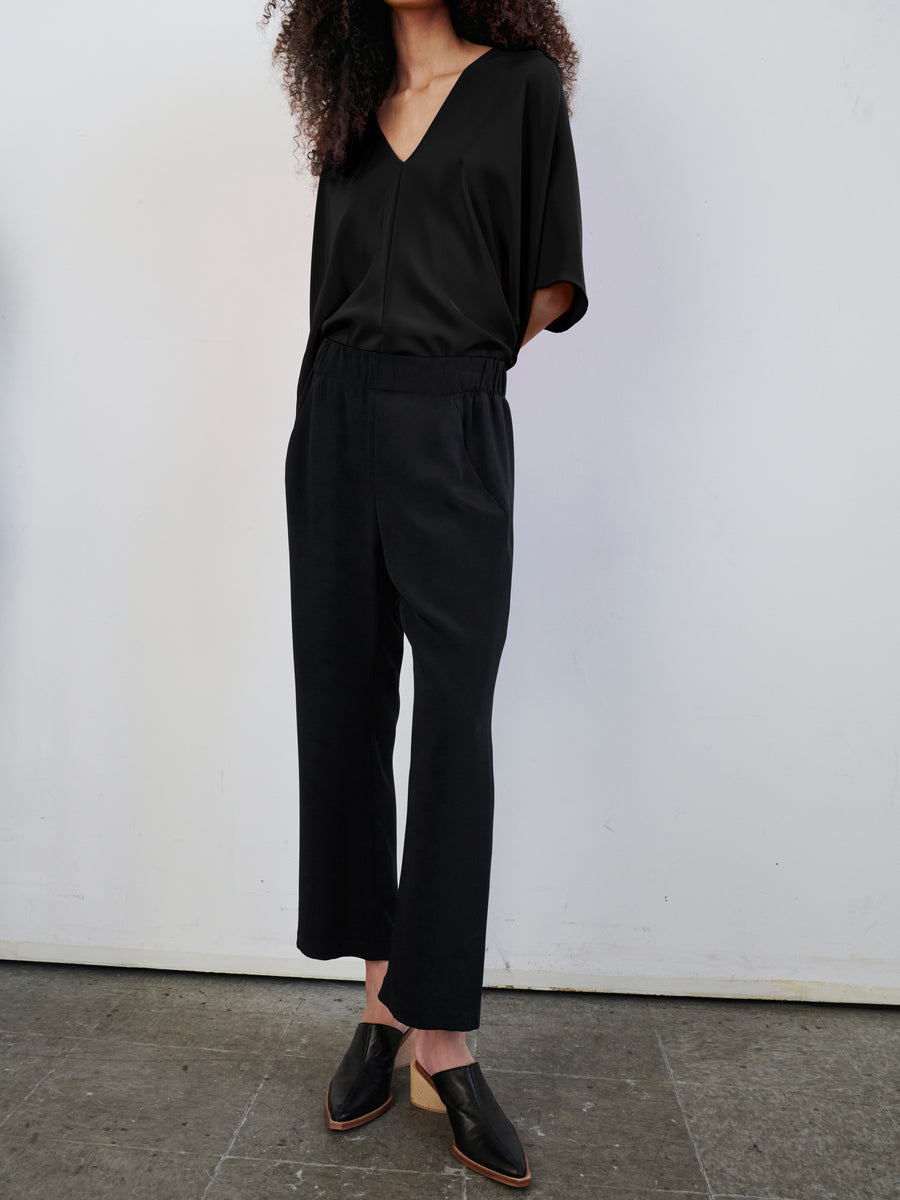 A person with curly hair is standing against a plain white wall, dressed in a loose black V-neck top and the Eko Pant by Zero + Maria Cornejo. The wide-leg woven silk trousers feature an elastic waist and hidden pockets. They complete the look with black slip-on shoes.