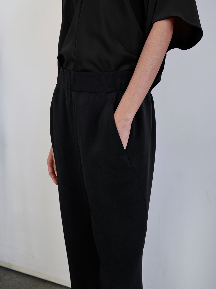 A person wearing a black outfit stands against a plain white wall. Their right hand rests in the hidden pocket of their woven silk Eko Pant by Zero + Maria Cornejo, while their left arm is relaxed by their side. The clothing has a loose fit and smooth texture.