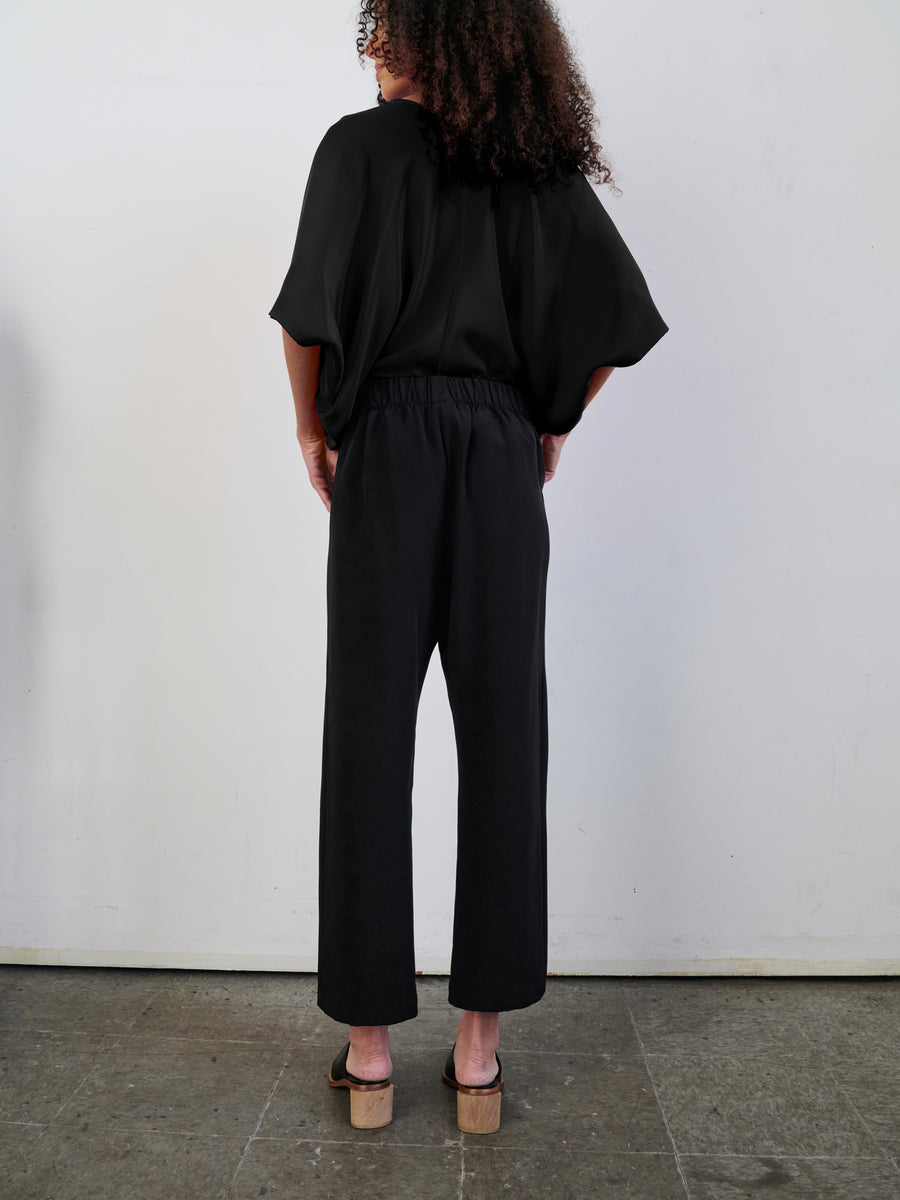 A person with curly hair is facing away, wearing a loose black top and the Eko Pant by Zero + Maria Cornejo, featuring woven silk and an elastic waist. They stand on a concrete floor against a plain white wall while wearing beige platform sandals. Hidden pockets enhance the effortless style of their ensemble.