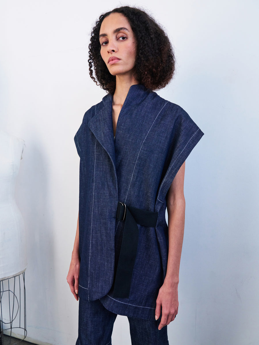 A person with curly hair is wearing the Ari Waistcoat, a sleeveless wrap vest in dark blue made from organic denim by Zero + Maria Cornejo, featuring a black belt. They stand against a plain wall, looking confidently forward.