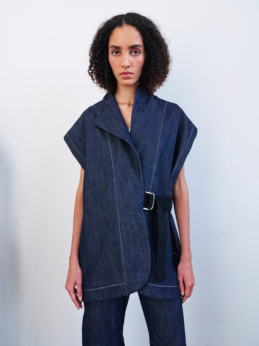 Against a white backdrop, a woman models the Ari Waistcoat by Zero + Maria Cornejo, showcasing its sleeveless, dark denim oversized design crafted from GOTS-certified cotton. Her curly hair frames her face as she accessorizes with a black belt and keeps a neutral expression.