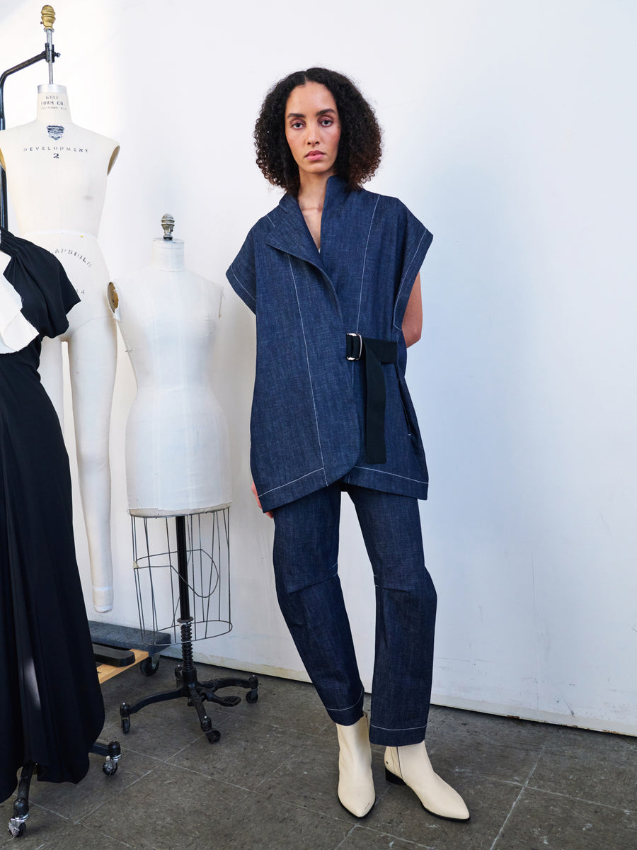 A person with curly hair dons the Ari Waistcoat, a sleeveless wrap jacket crafted from organic denim by Zero + Maria Cornejo, paired with matching pants. They stand beside dress forms, completing the look with white ankle boots against a plain white wall backdrop.