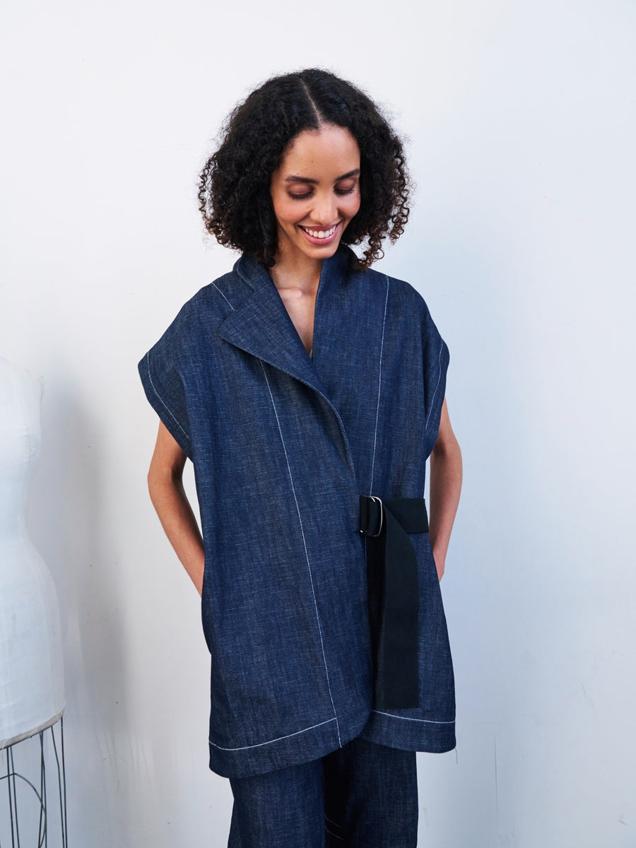 A person with curly hair smiles while looking down, wearing the Ari Waistcoat by Zero + Maria Cornejo. This loose-fitting, organic denim waistcoat features a wide collar and belt against a simple white background.