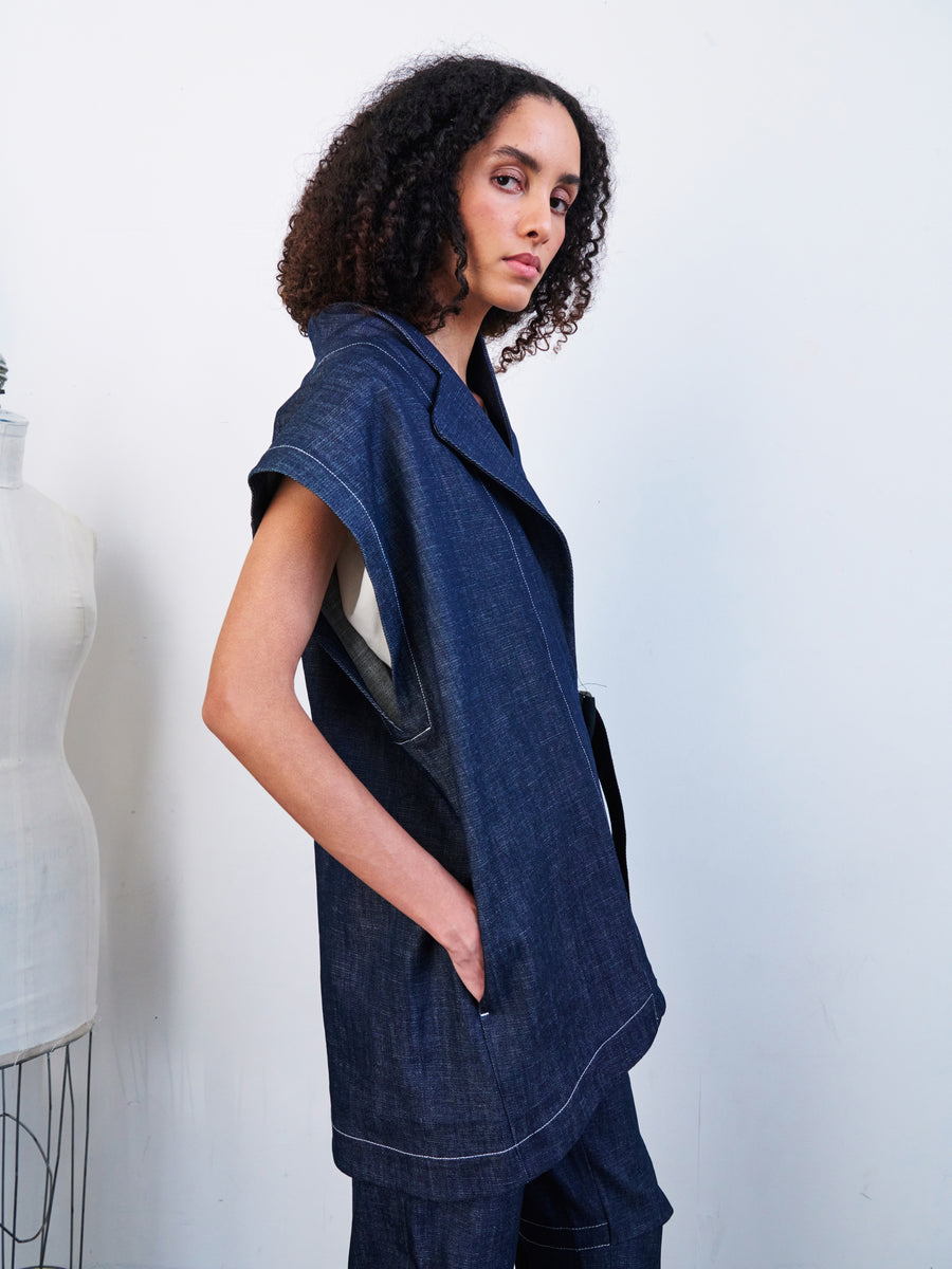 A person with curly hair is wearing the Ari Waistcoat by Zero + Maria Cornejo, along with matching denim pants. The pose is sideways, with one hand in the waistcoat pocket, set against a plain white background next to a dress form. The outfit's sustainable design reflects eco-friendly fashion choices.